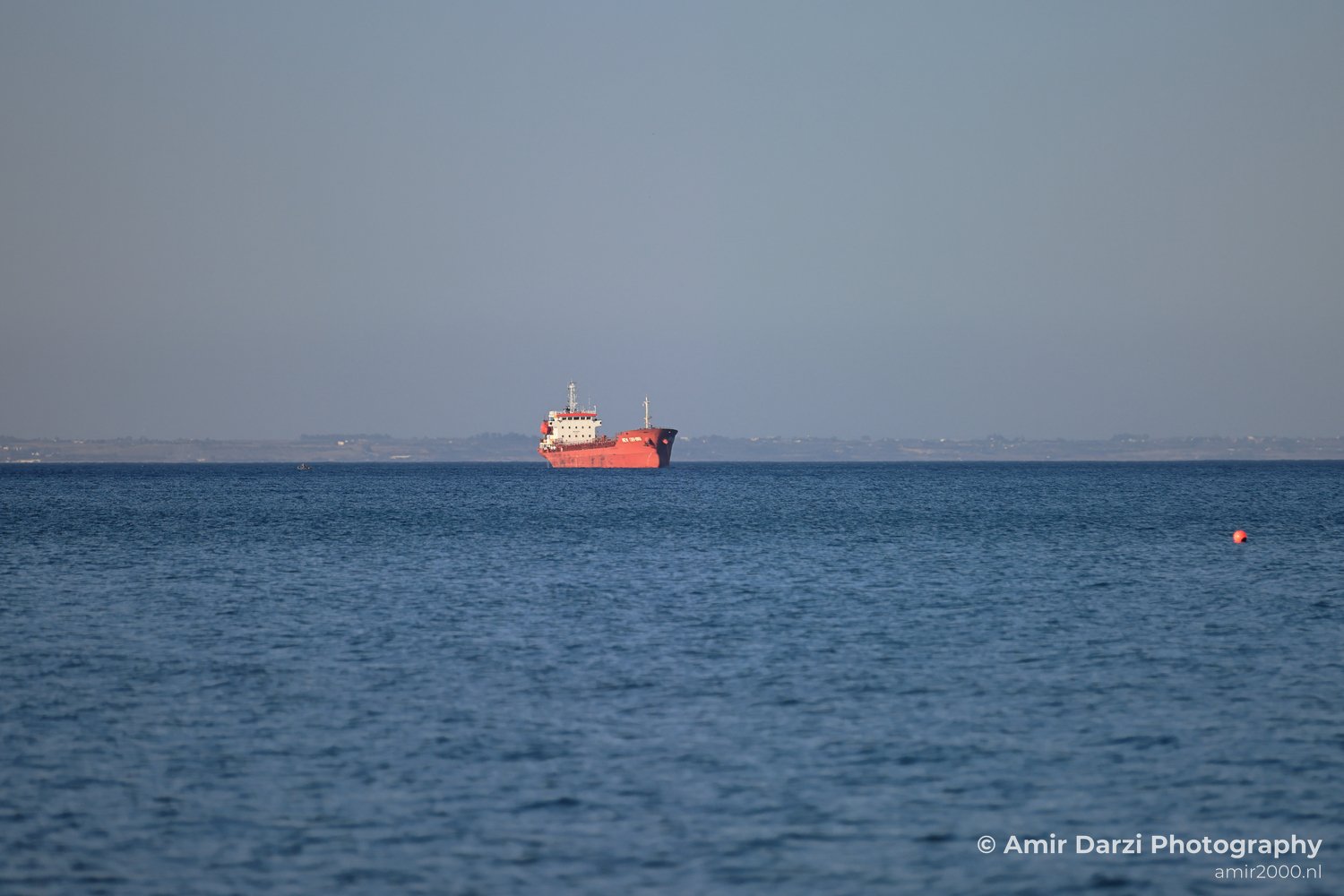 Red cargo ship is positioned centrally on calm blue waters under a clear sky, with land faintly visible in the distance in Larnaca, Cyprus.