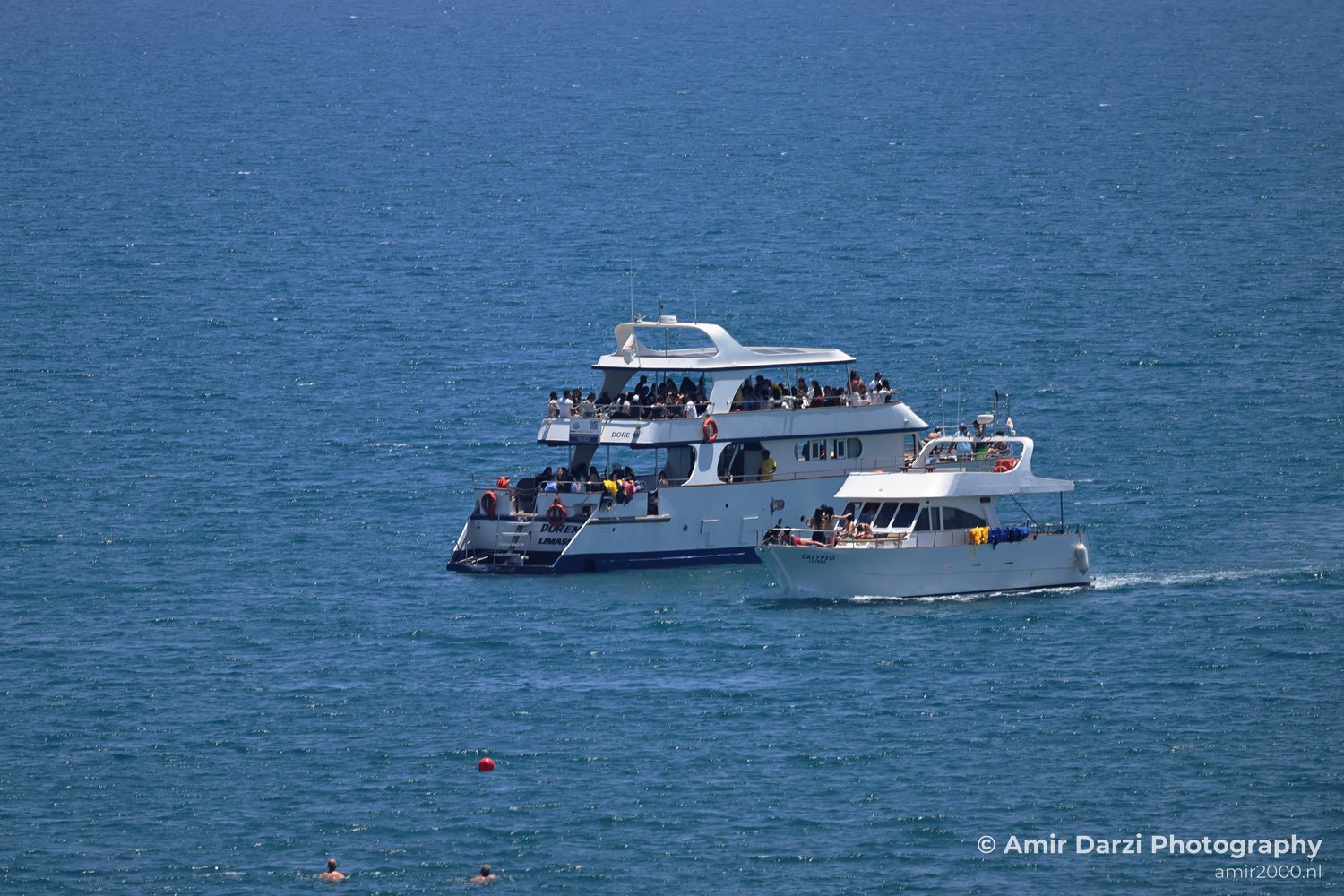 A vast blue sea, surrounded by smaller boats and people enjoying the water near it includes the large white boat named "CALYPSO" sailing.