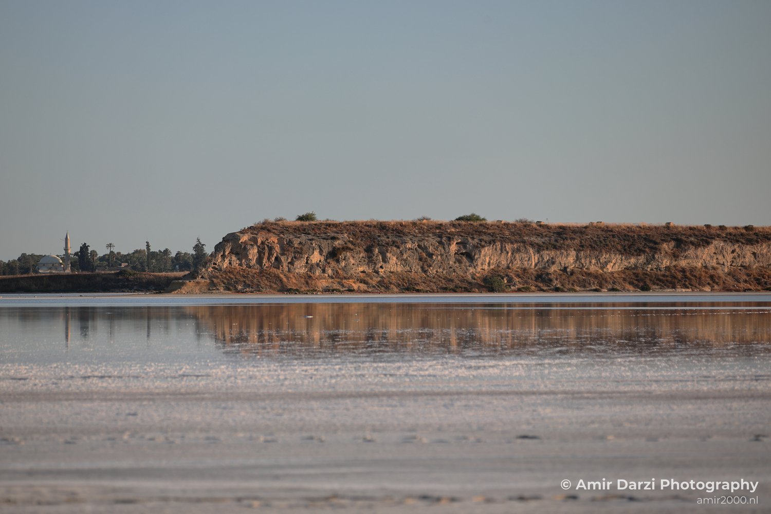 A saltwater body reflecting the sky and a distant rocky outcrop, with a mosque visible along the shoreline.