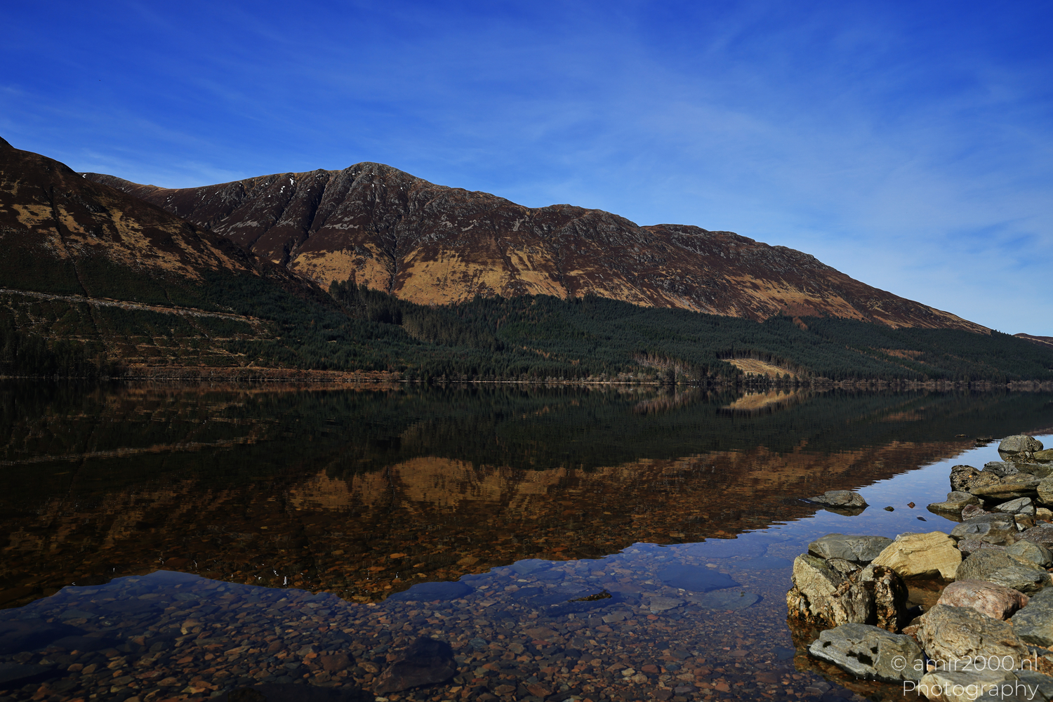Rocky Loch Lochy shore reflecting mountains and trees in Scotland