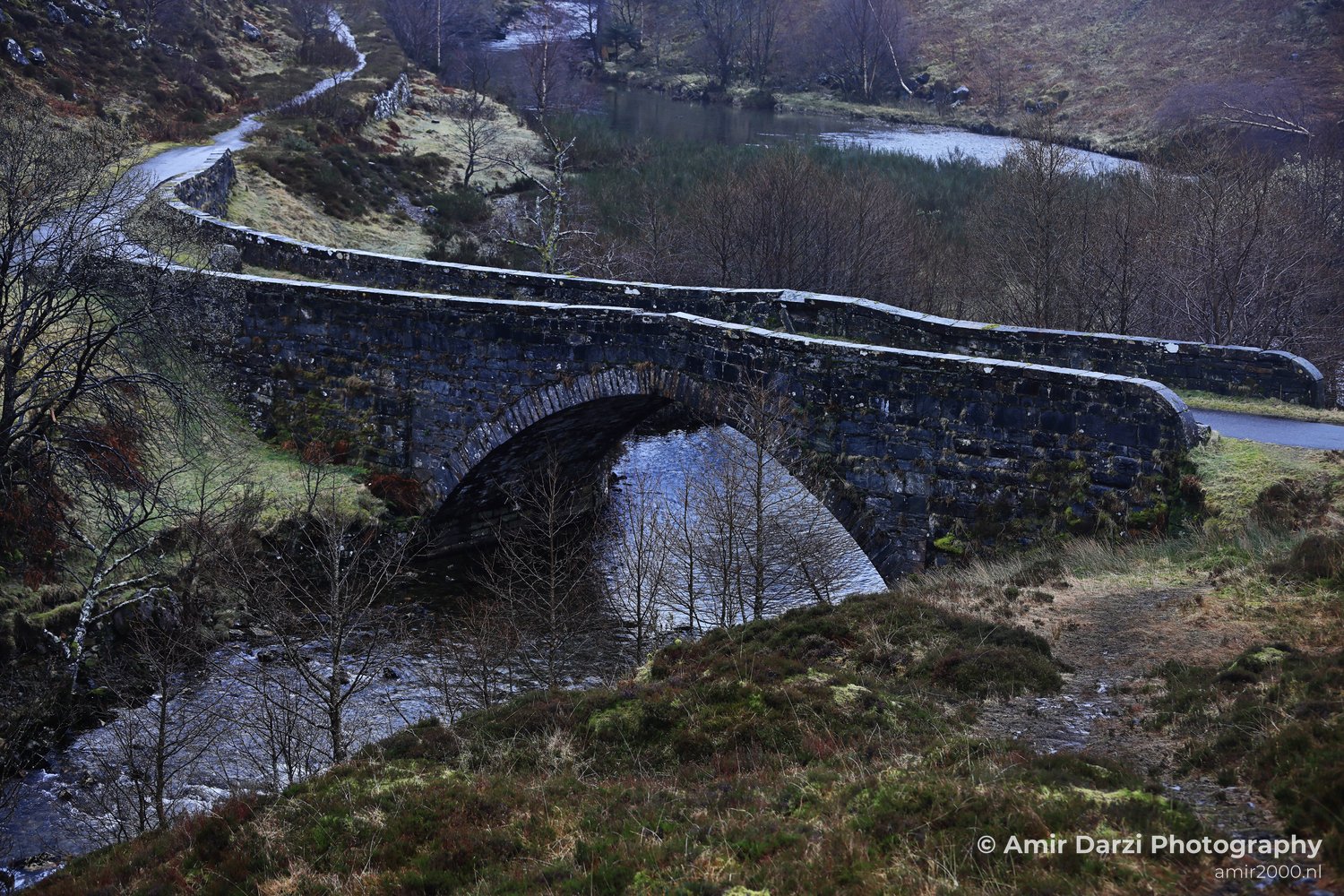 Bare trees and grassy banks surrounding it appear with the stone bridge spanning over a river in Scotland United Kingdom.