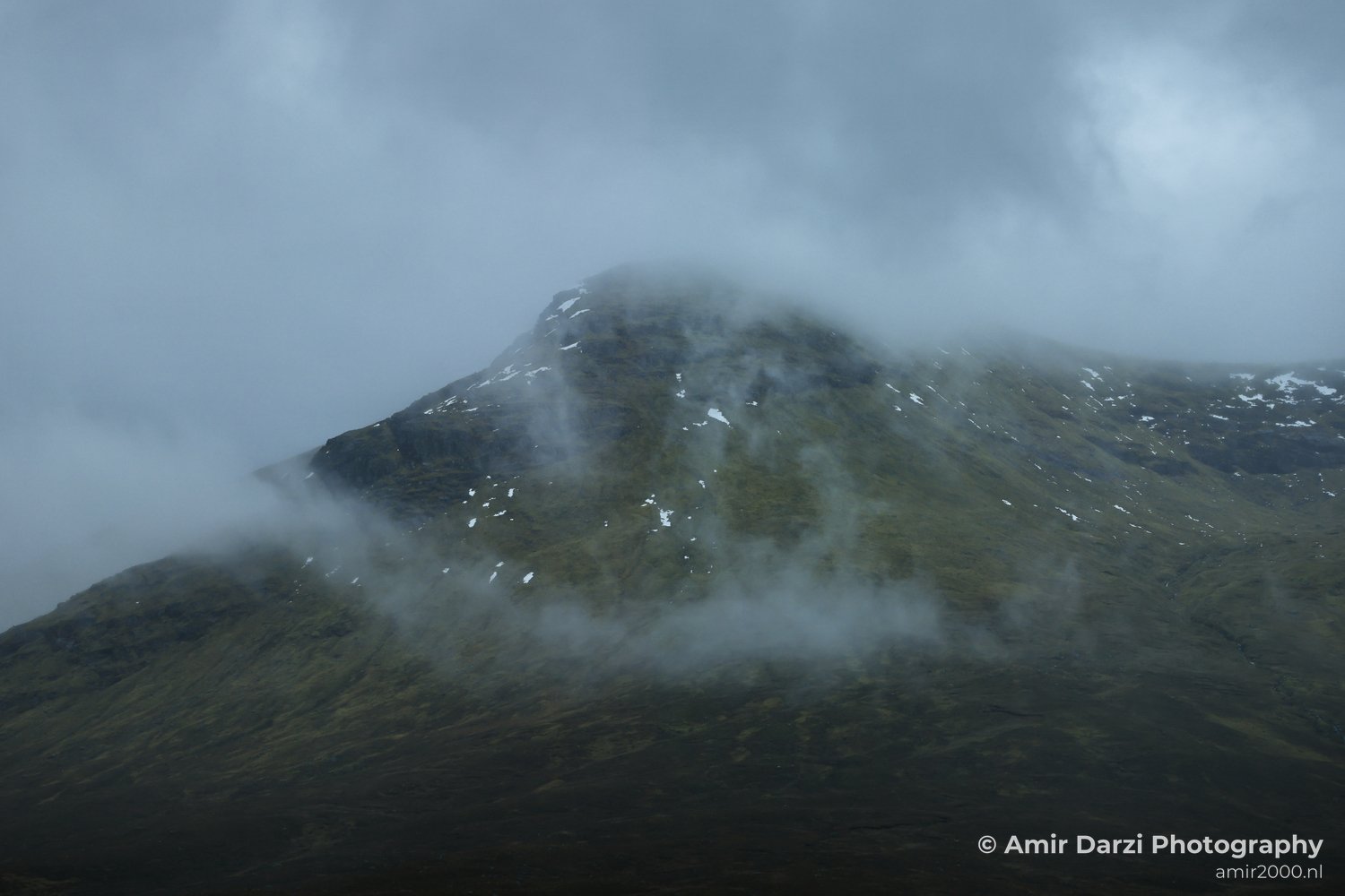 Patches of snow and grass, partially obscured by clouds and mist, appear with the mountain covered in Scotland United Kingdom.