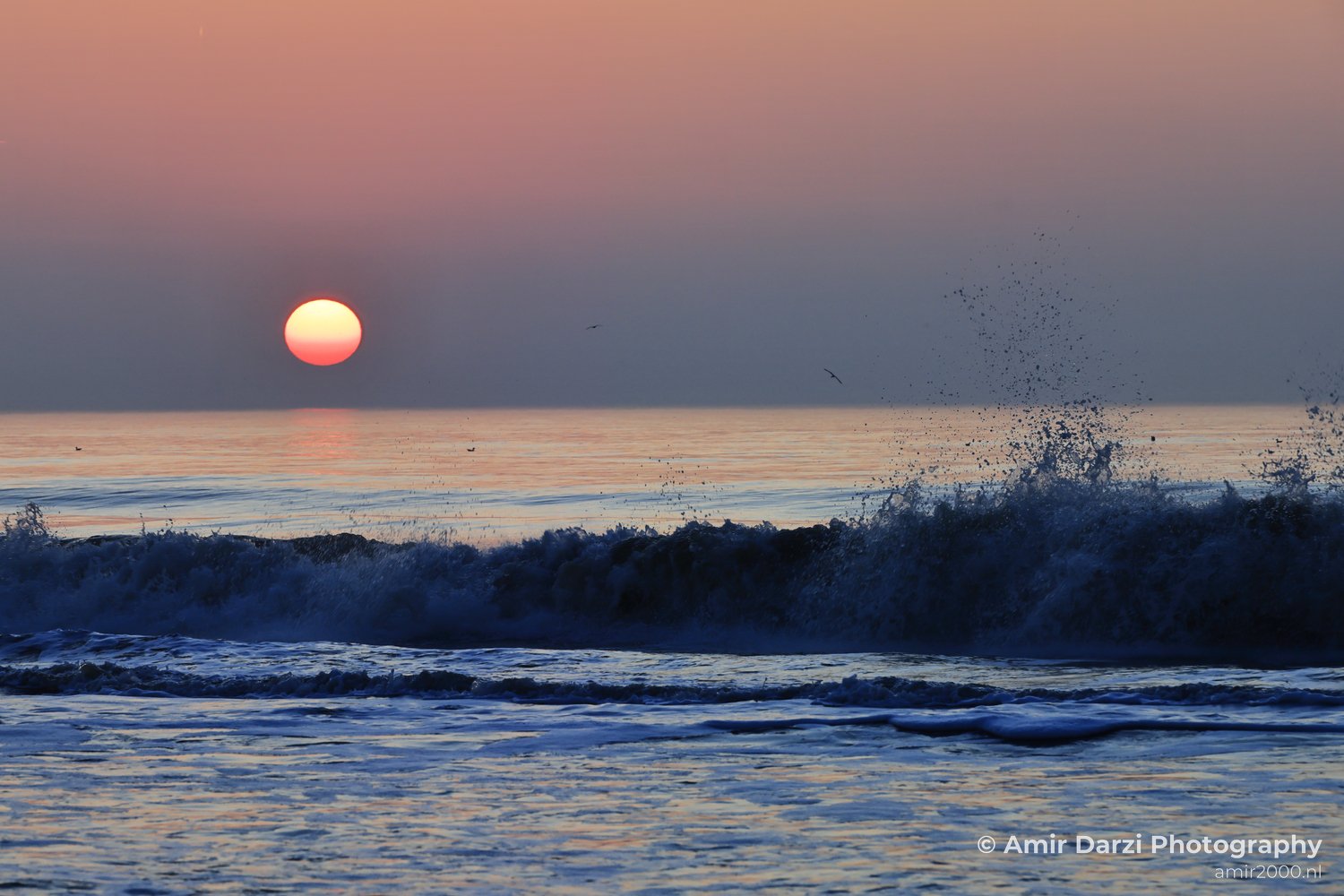 Waves break along the North Sea shoreline under a warm sunset sky.