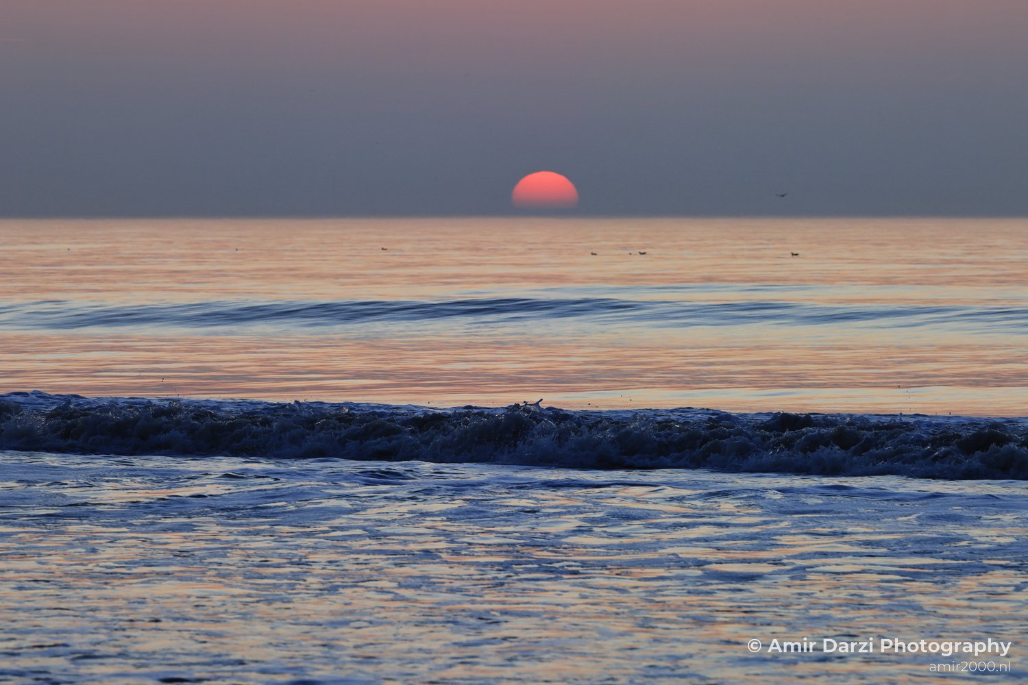A sunset over the North Sea, with the sun setting low and waves gently lapping against the shore.