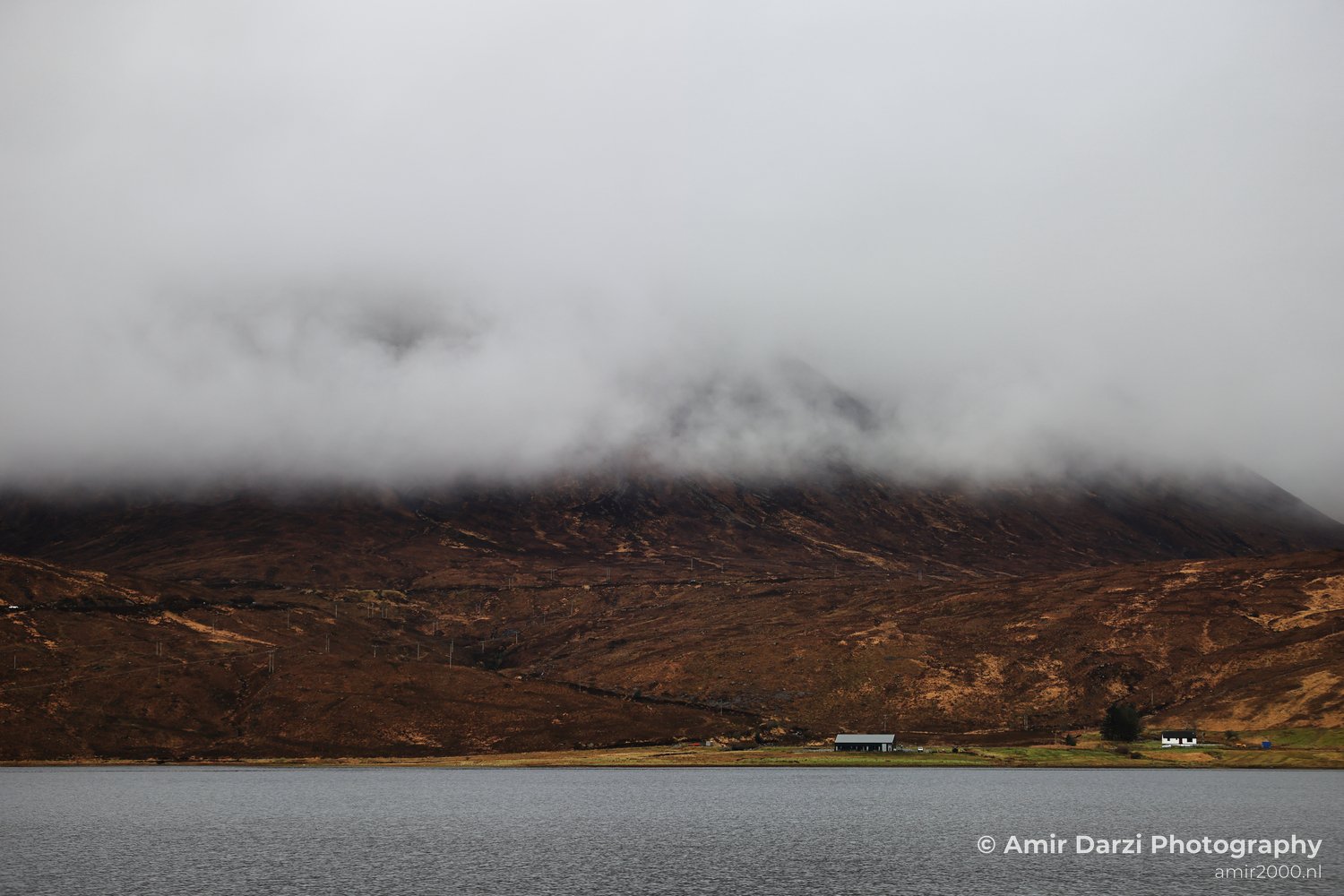 A body of water in the foreground, a grassy area with some structures near it, and a mountain covered partially by clouds in the background, appears with the landscape in Scotland United Kingdom.