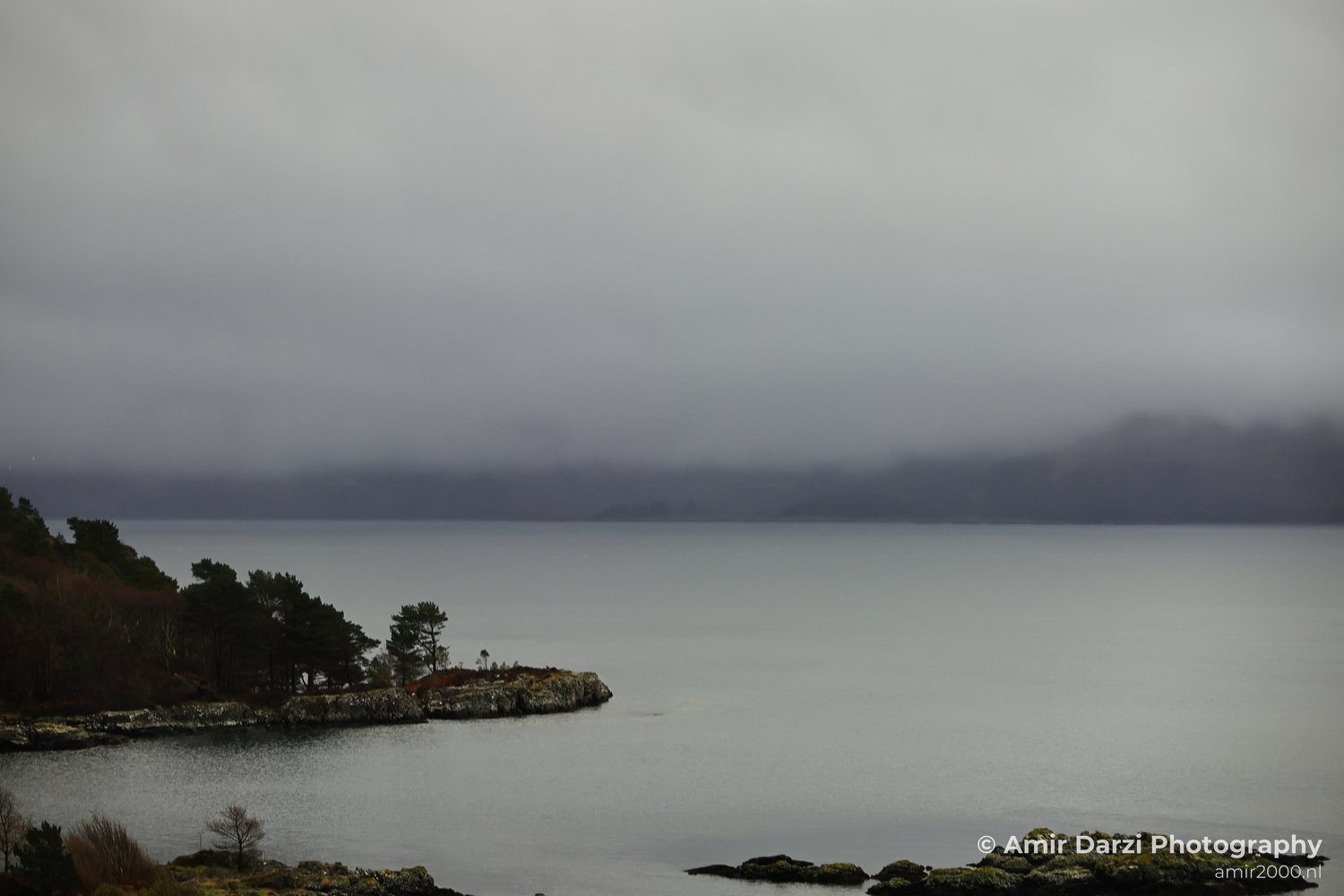 Calm water, rocky shorelines, sparse vegetation, and an overcast sky, appears with the serene coastal scene in Scotland United Kingdom.