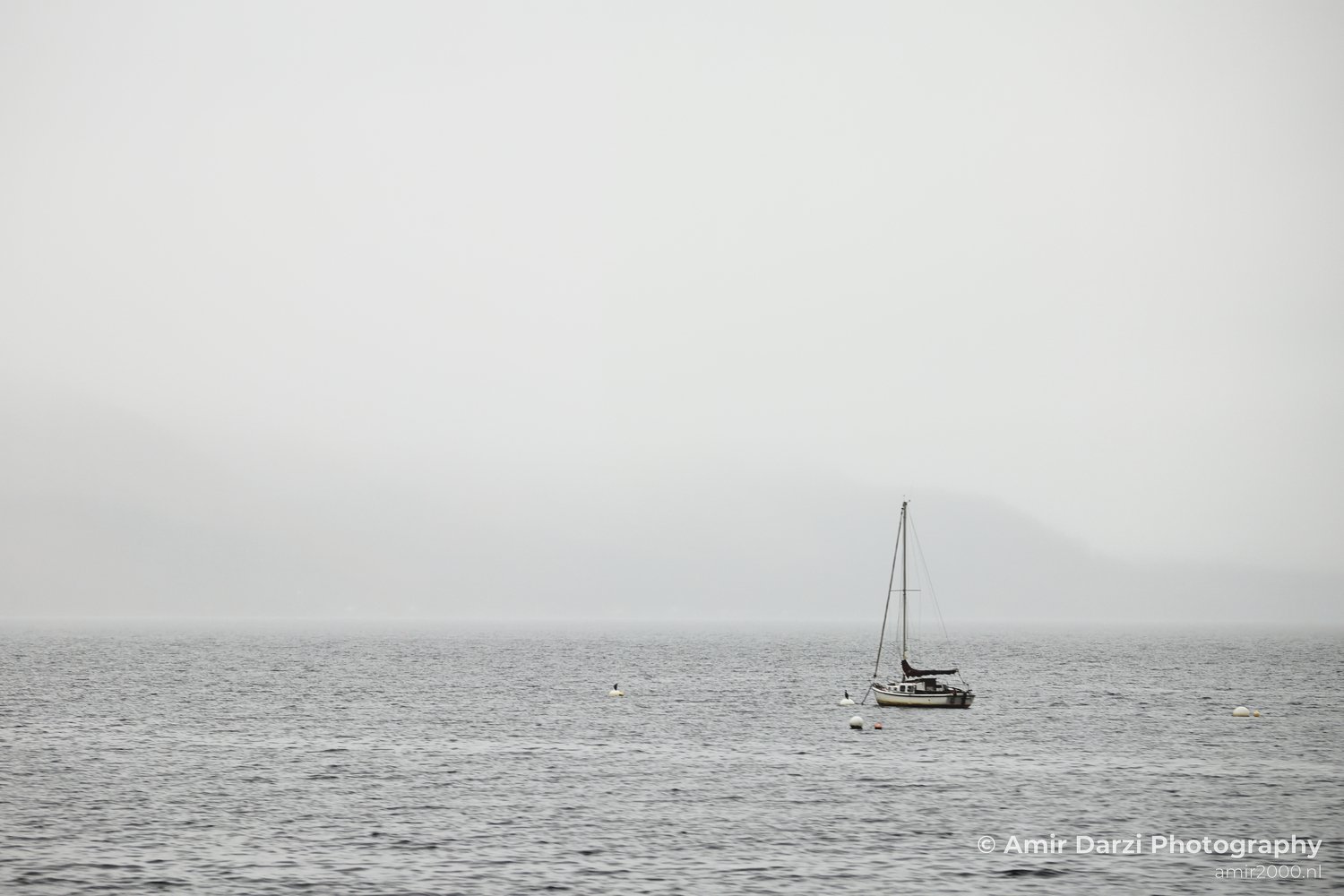 Some buoys and a foggy horizon appears near the calm body of water with a single sailboat in Scotland United Kingdom.