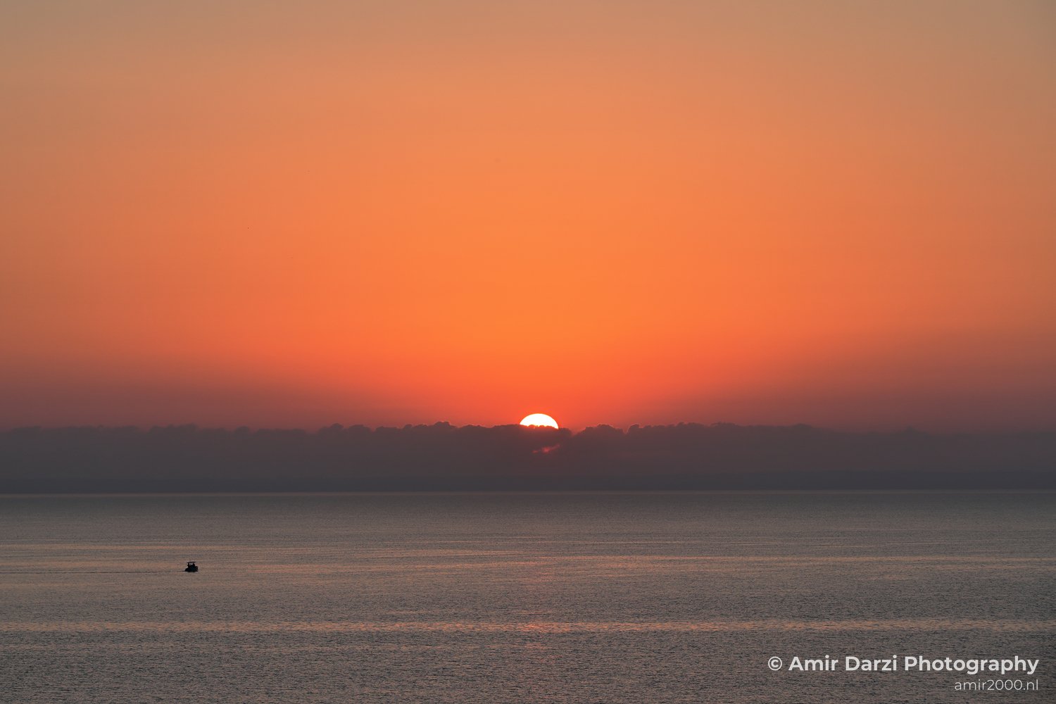 The sun partially obscured by clouds, casting an orange hue across the sky, appears alongside a scene where a serene sunset over calm waters where a small boat is seen near the horizon in Larnaca, Cyprus.
