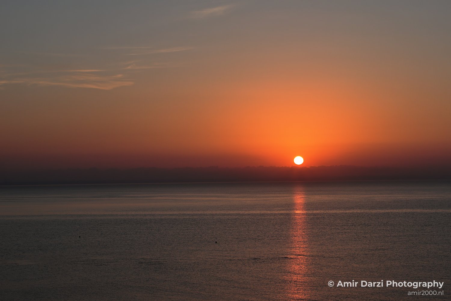 Subtle clouds above and reflections on the surface below appear alongside a scene where the sun is setting over calm water, casting an orange glow across the sky in Larnaca, Cyprus.