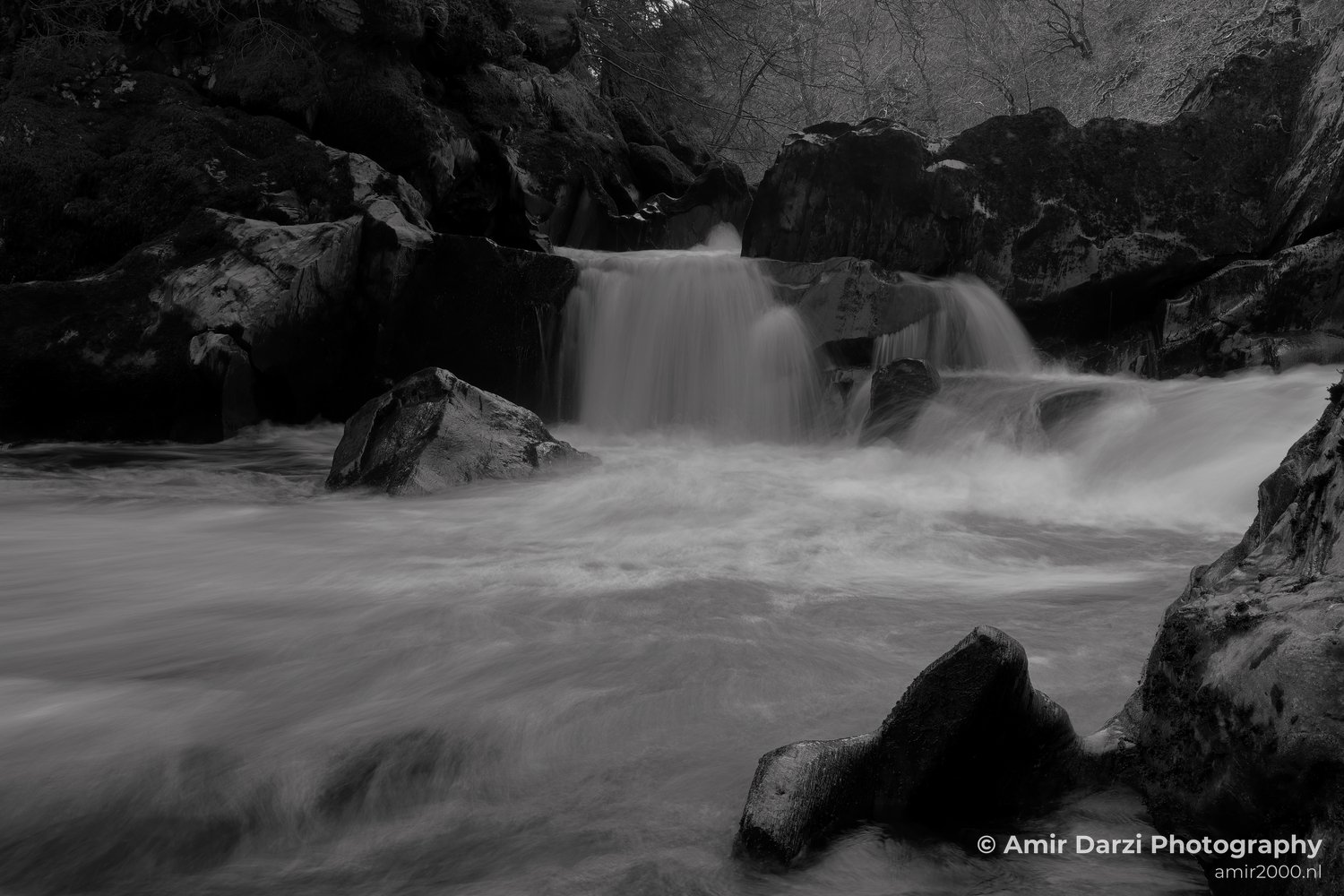 Rocks into a flowing river surrounded by more rocky terrain and trees includes the black and white image of a waterfall cascading.