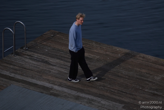 A young man in a blue sweater and black pants stands on a wooden dock near the water. image from year 2026 #2