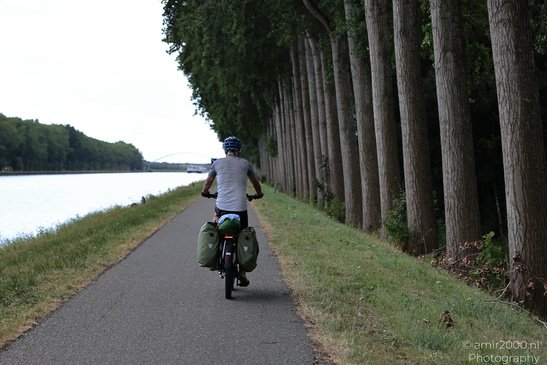 A person wearing a helmet rides a bicycle along a paved path beside a river, with tall trees - image from year 2026 #001