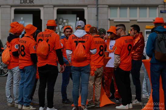 Group of people wearing orange jerseys and hats, standing near a street with buildings and a - image from year 2026 #011