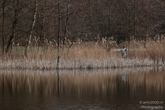 Person standing near a body of water with reeds and trees in the background in Naarden, - image from year 2026 #002