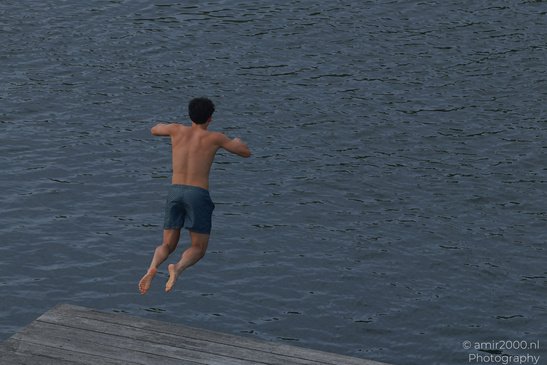 A shirtless person wearing patterned shorts is mid-air, leaping off a wooden dock into a water - image from year 2026 #001