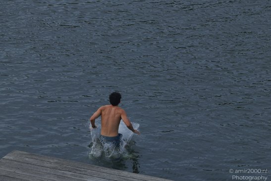 A shirtless person is running into a body of water, creating splashes, with a wooden dock in the - image from year 2026 #003