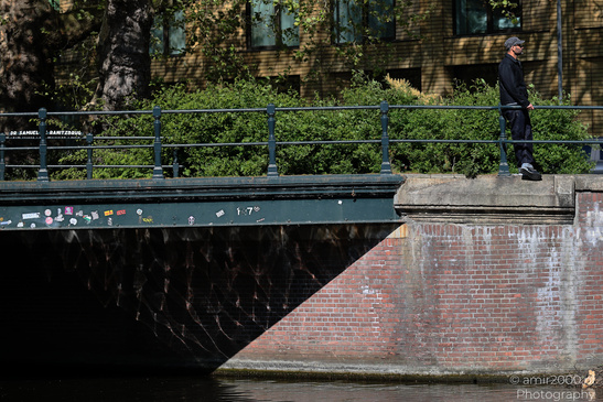 A lone figure stands on a bridge over a canal, with graffiti and brickwork visible. - image from year 2026 #001