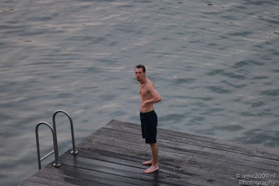 A shirtless man in dark shorts stands barefoot on a wooden dock by a body of water, with a metal - image from year 2026 #001