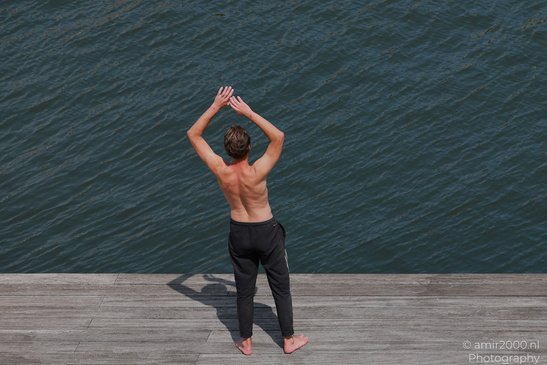 A shirtless individual wearing black pants stands on a wooden dock, raising their arms above - image from year 2026 #002