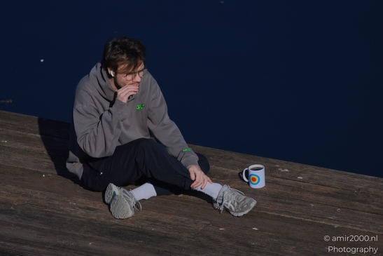Man sitting with coffee on a wooden dock beside dark canal water in Amsterdam Netherlands. - image from year 2026 #001
