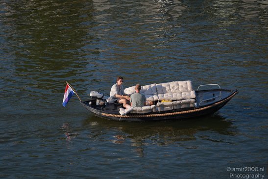 Two people relax on a boat with a Dutch flag, surrounded by calm water in Amsterdam, Netherlands. - image from year 2026 #007