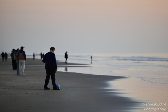 A solitary figure on a beach at sunset, kicking a soccer ball, with the horizon and distant figures. image from year 2026 #3