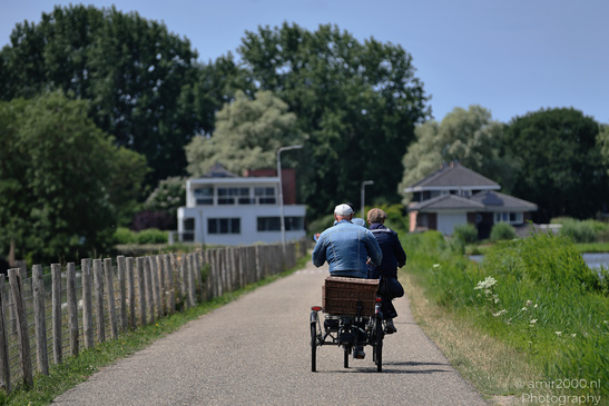 Rear view of two people riding a tricycle on a paved path between trees and fields. image from year 2026 #1