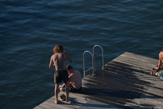 Two shirtless individuals with tattoos on a wooden dock by a body of water, with one person - image from year 2026 #001