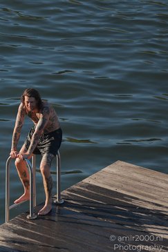 A tattooed individual wearing black swim trunks is standing on a wooden dock, holding onto a - image from year 2026 #005