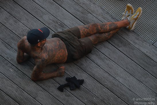 A tattooed man lies on a wooden dock, wearing a cap and sneakers, with a sock in Amsterdam, - image from year 2026 #001