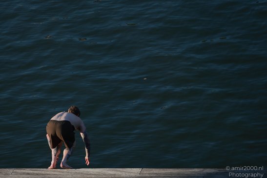 A person with tattoos on their arms and legs is bending over near a body of water, standing on a - image from year 2026 #001