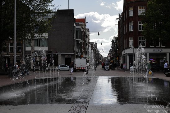 A street scene with a fountain in the foreground, buildings on either side, a white van parked, - image from year 2026 #001