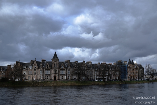 A row of stone buildings with turrets and arched windows along a riverbank under an overcast sky - image from year 2026 #006