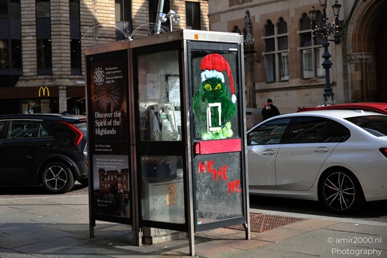 A public telephone booth with a painted Grinch character wearing a Santa hat and holding a sign - image from year 2026 #009