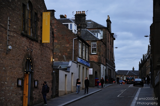 A narrow street in a Scottish city at dusk, with people walking and buildings lining the sides. - image from year 2026 #019