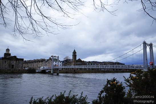 A Scottish cityscape with a suspension bridge over a calm river and historic buildings. - image from year 2026 #020