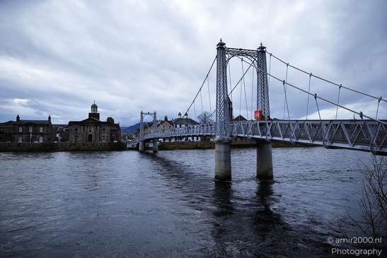 Scottish City Inverness a suspension bridge spans a calm river with buildings and trees. - image from year 2026 #021