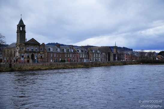 A Scottish cityscape with a church and houses along the riverbank under an overcast sky. - image from year 2026 #025