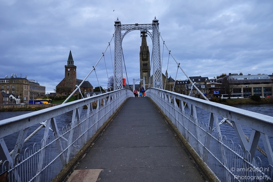 A suspension bridge in a Scottish city with a church and clock tower. - image from year 2026 #028