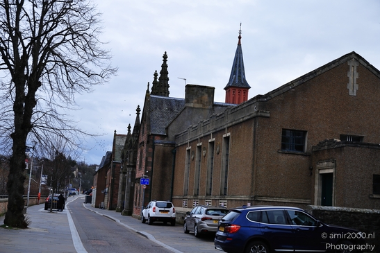 A quaint Scottish town street with a historic church and modern cars parked along the side. - image from year 2026 #030