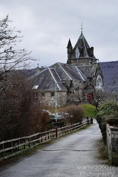 A stone building with a steeply pitched roof and a tower featuring a weathervane, surrounded by - image from year 2026 #005