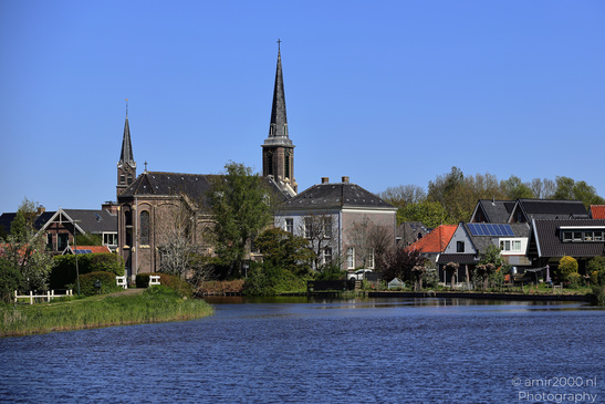 A village by a calm lake with historic buildings and a clear blue sky. - image from year 2026 #001