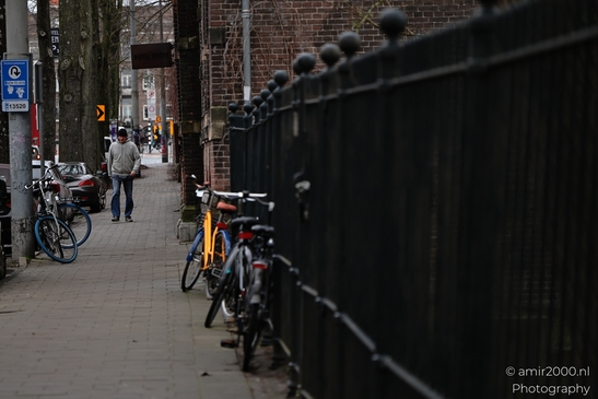 A row of bicycles parked against a fence with pedestrians walking nearby and brick buildings lining. image from year 2026 #1