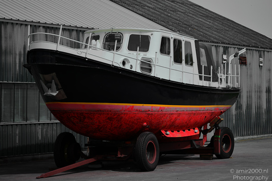 A boat with a black hull and red bottom section on wheels, parked outside near a corrugated - image from year 2026 #001