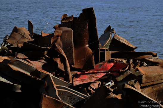 A pile of rusted metal debris with some pieces having red paint on them, situated near a body of - image from year 2026 #002