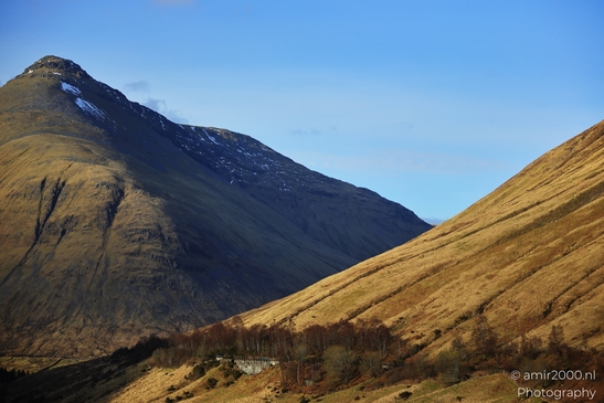 A rugged mountain landscape under a clear blue sky, showcasing steep slopes covered in dry grass - image from year 2026 #017