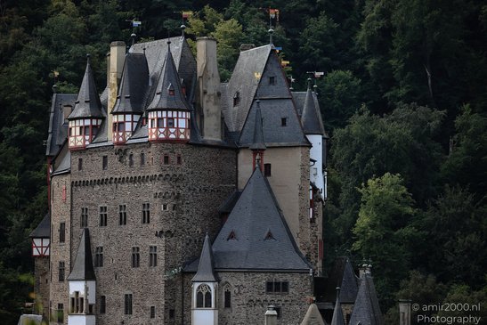 A medieval castle with gray stone walls and pointed roofs, surrounded by lush green trees in - image from year 2026 #008