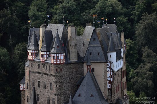 A medieval castle with dark gray rooftops and stone walls, surrounded by lush green trees in - image from year 2026 #012