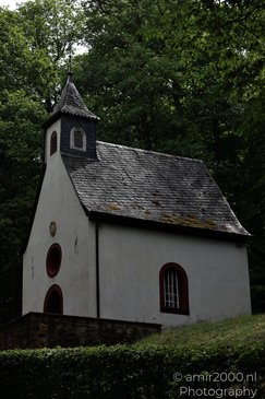 A small white chapel with a dark tiled roof, surrounded by trees and greenery in Rhineland - image from year 2026 #013
