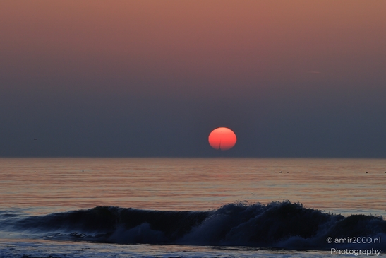 A sunset over the North Sea, with a red sun setting behind the horizon, and gentle waves. - image from year 2026 #017